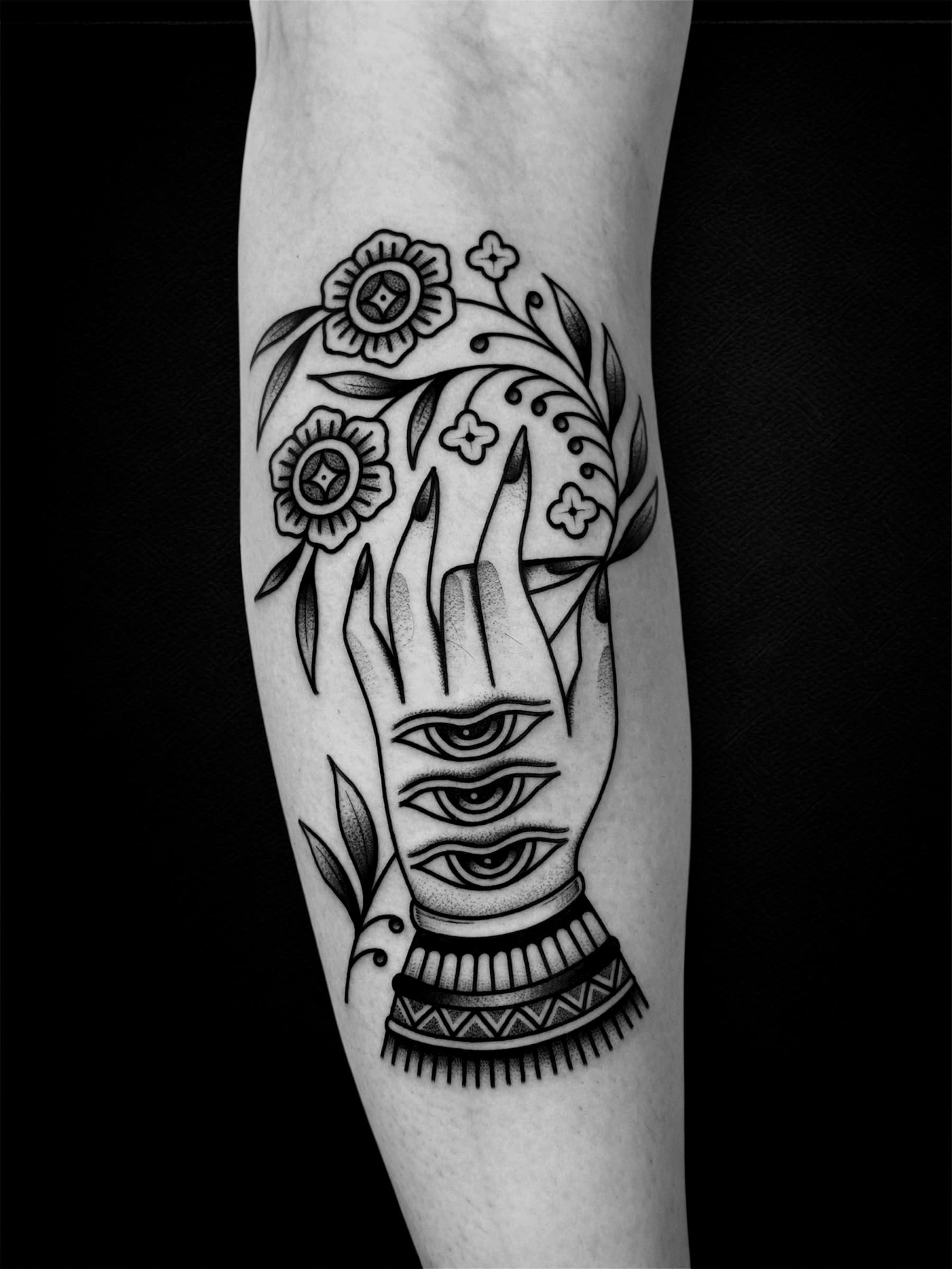 Tatouage ornemental noir et gris d’une main aux trois yeux entourée de fleurs